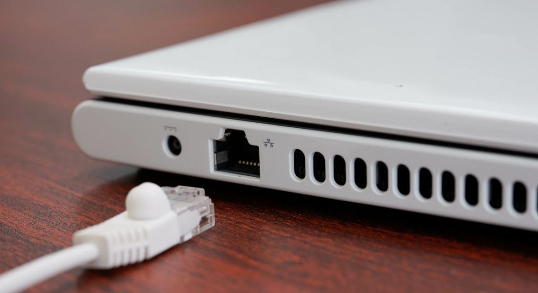 A white ethernet cable lies beside its port on a white laptop on a dark brown desk.