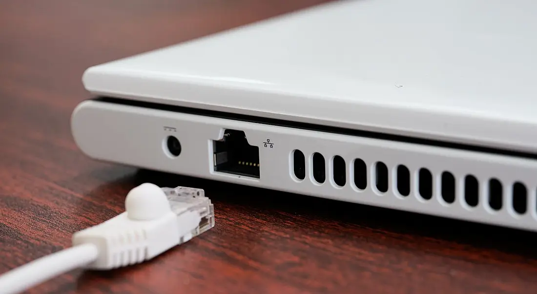 A white ethernet cable lies beside its port on a white laptop on a dark brown desk.