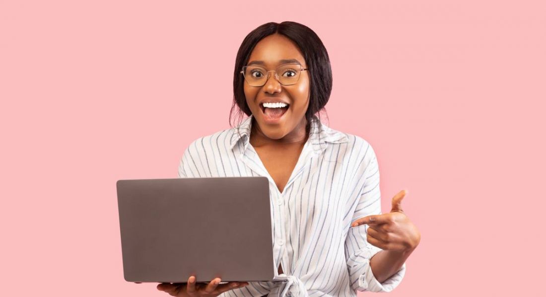 A woman is standing against a pink background and pointing at her laptop excitedly before applying for new jobs.