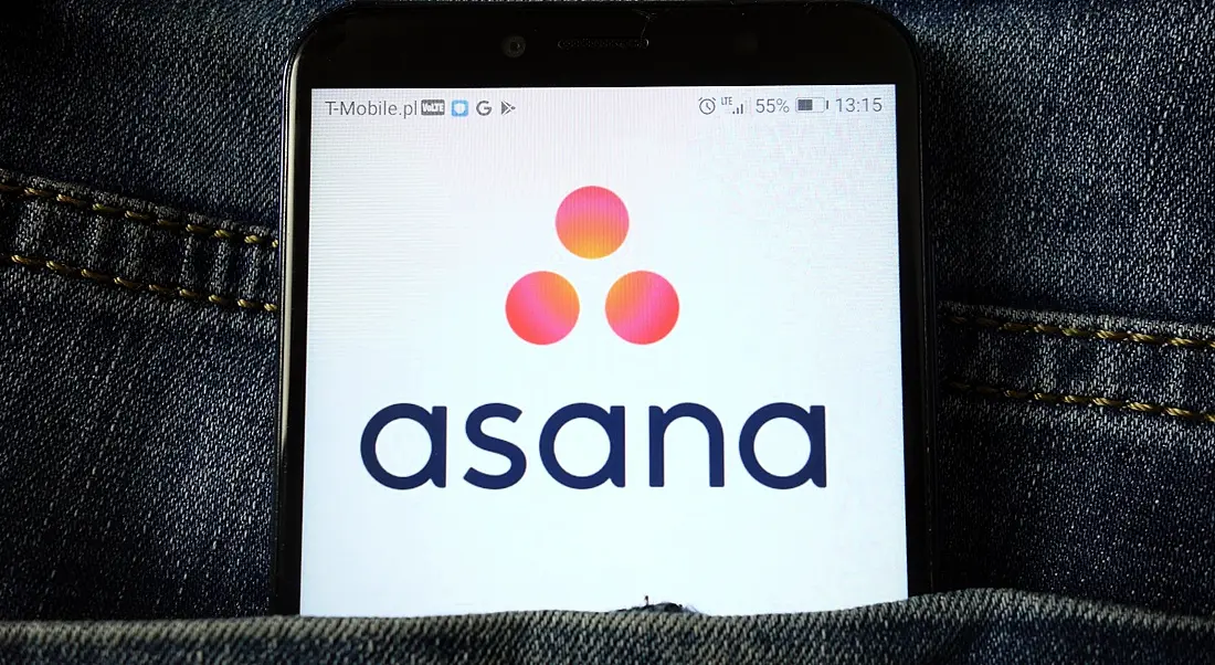 The Asana app logo is displayed on a mobile phone, which is stick out of a person's trouser pocket. The logo includes the name of the company and three pink dots.