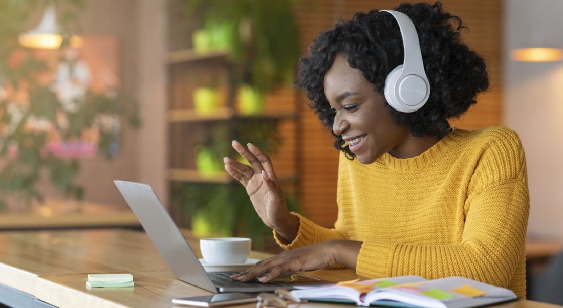 A young woman is sitting in front of her laptop in a cafe, wearing headphones and speaking to someone on a video conference call.