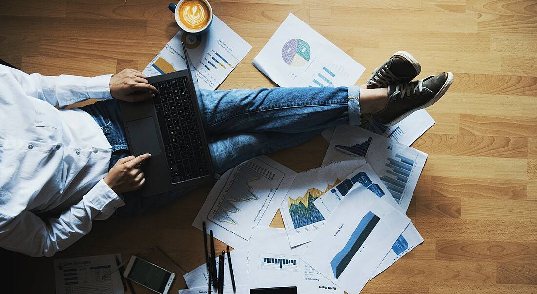 A woman is sitting on a wooden floor surrounded by papers with a laptop while working from home.