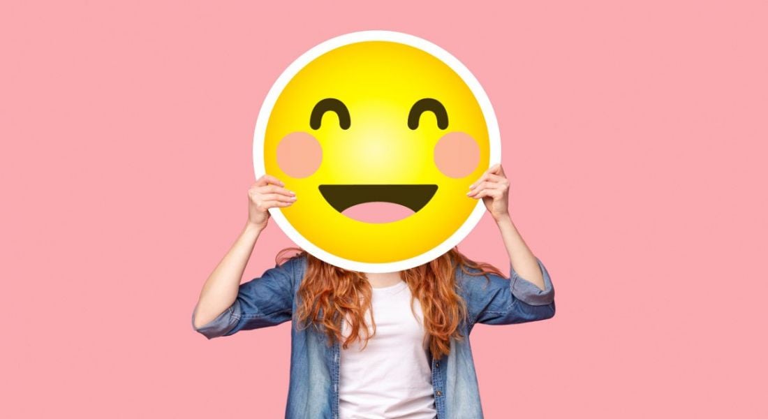 A woman in casual clothing is holding a large image of a smiling emoji above her face.