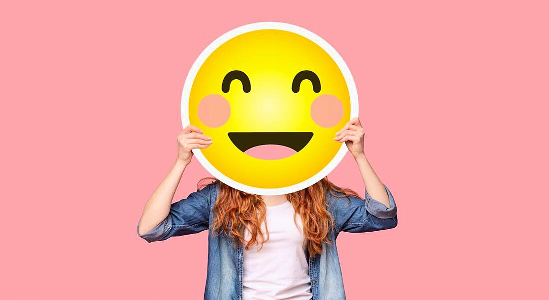 A woman in casual clothing is holding a large image of a smiling emoji above her face.