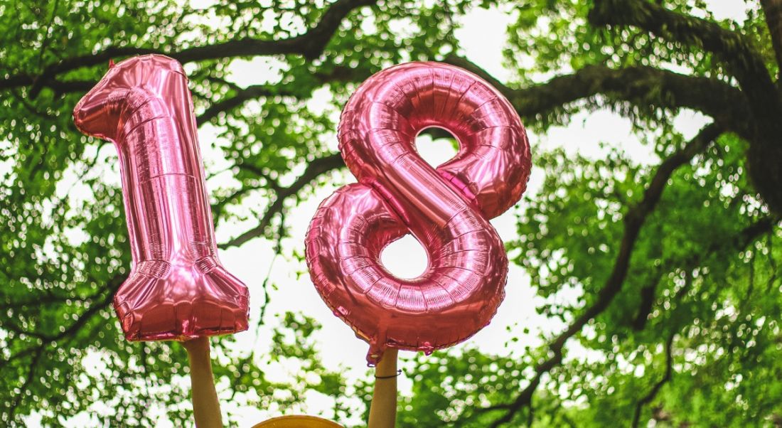 Pink foil number 18 balloons are being held up against trees with leaves and a blue sky.