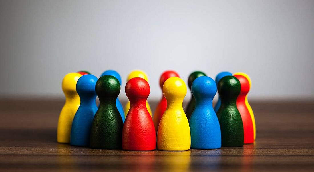 Wooden figures of all different colours huddled together on a wooden desk, representing diversity and inclusion.