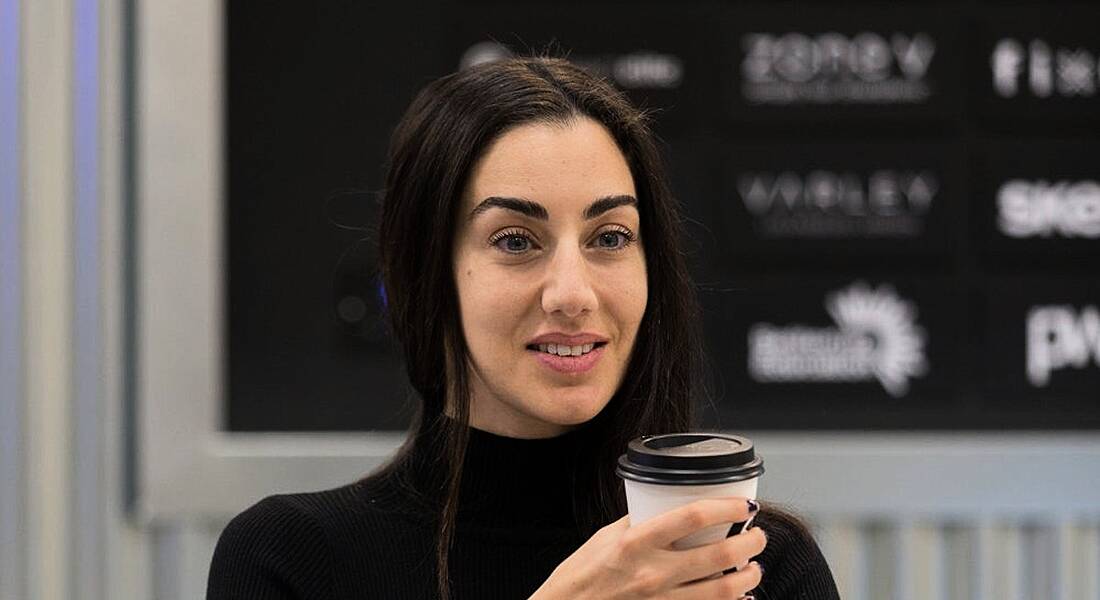 A woman with dark hair wearing a black polo neck jumper, holding a takeaway coffee cup and smiling off camera.