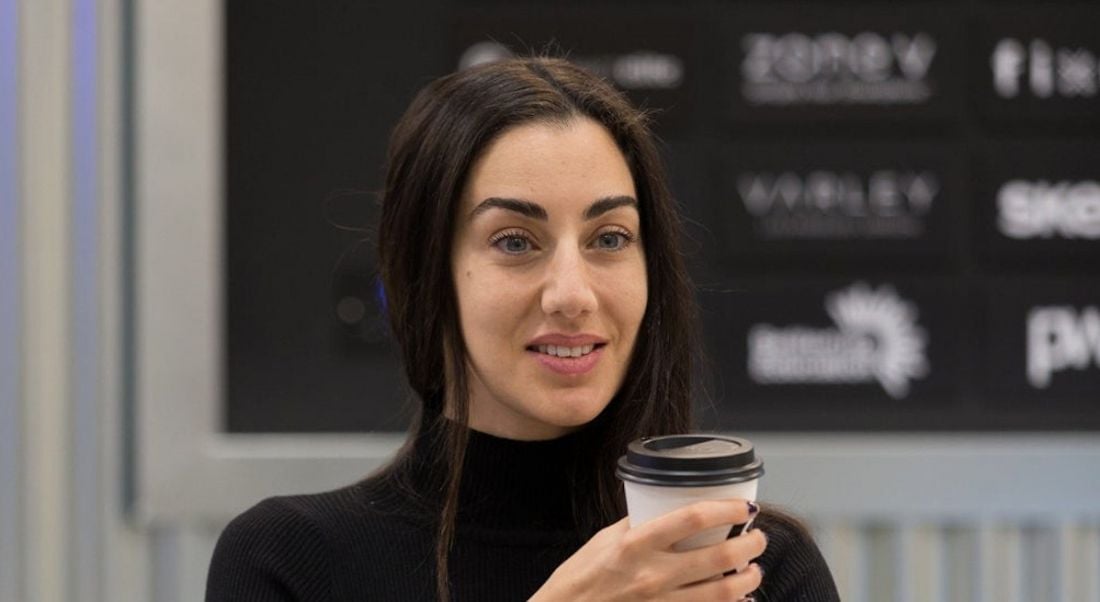 A woman with dark hair wearing a black polo neck jumper, holding a takeaway coffee cup and smiling off camera.