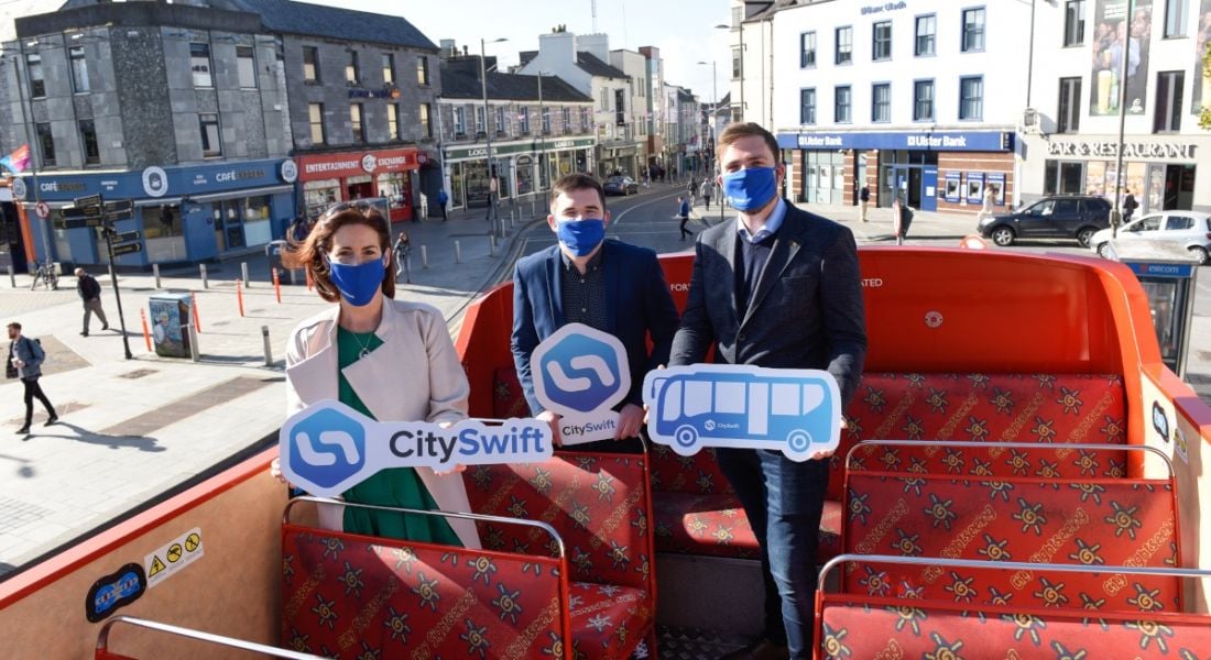 A woman and two men sit on an open-top red bus in Galway city. They're all wearing blue face coverings and holding signs with the CitySwift logo or a picture of a bus.