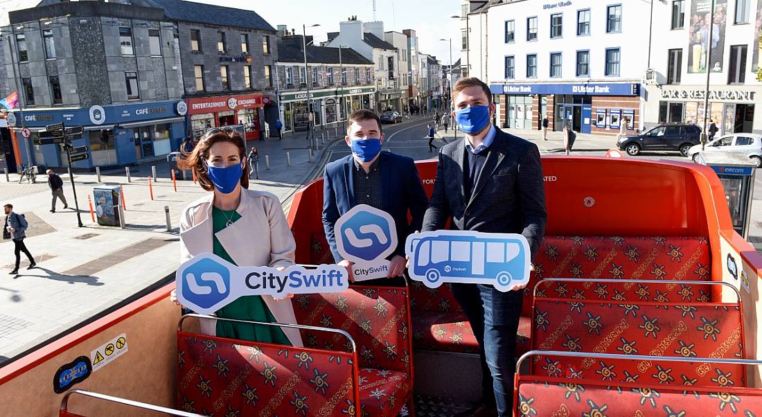 A woman and two men sit on an open-top red bus in Galway city. They're all wearing blue face coverings and holding signs with the CitySwift logo or a picture of a bus.