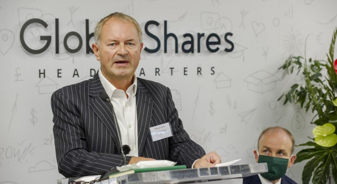 Tim Houstoun stands at a podium in front of a sign that reads 'Global Shares', with Taoiseach Michael Martin sitting in the background.