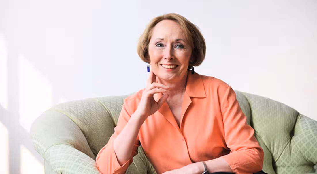 Expert on imposter syndrome Dr Valerie Young is sitting on a green couch in a brightly lit room and smiling into the camera.