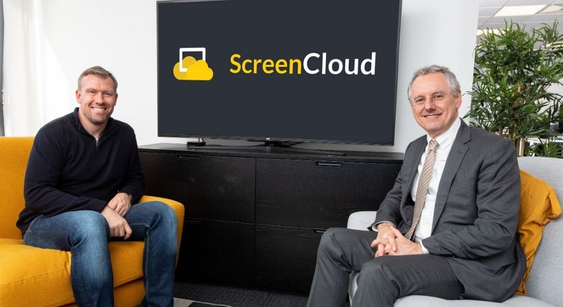 The ScreenCloud CEO is sitting beside the CEO of InvestNI in a modern office space in front of a widescreen TV with the company logo on it.