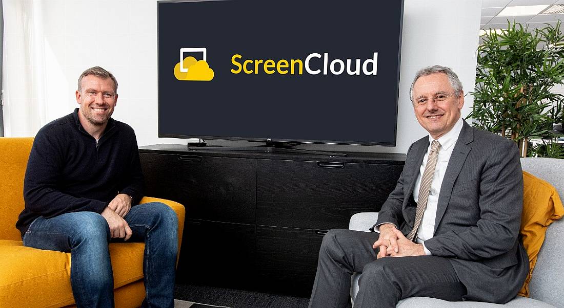 The ScreenCloud CEO is sitting beside the CEO of InvestNI in a modern office space in front of a widescreen TV with the company logo on it.