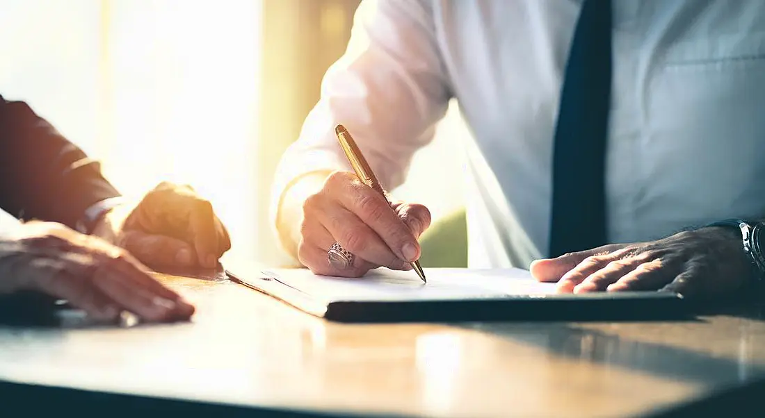 Close-up of a businessman signing a contract on a table with a pen as another businessman looks on.