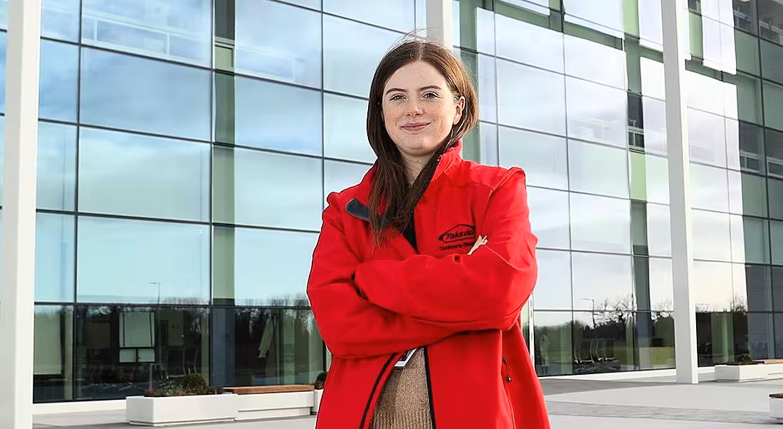 A young professional woman in casual clothing is standing outside the Takeda Dunboyne building, and wearing a red Takeda-branded jacket.