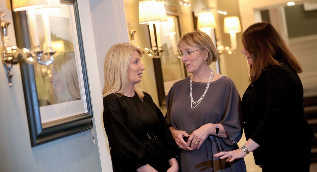 Three women stand in conversation in a softly lit hallway with artwork hanging on the walls.
