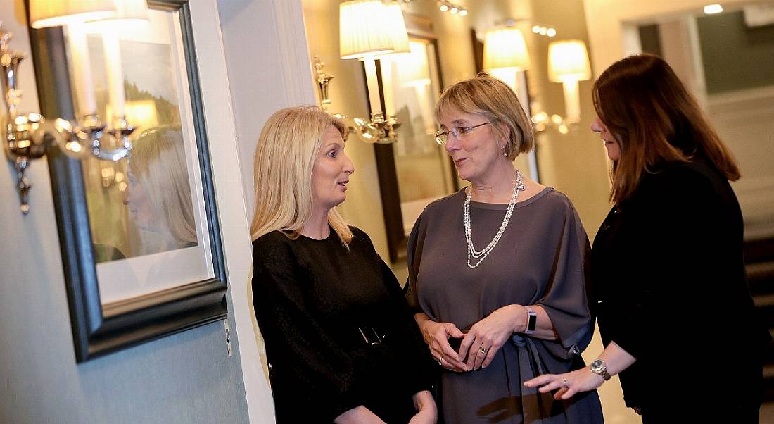 Three women stand in conversation in a softly lit hallway with artwork hanging on the walls.