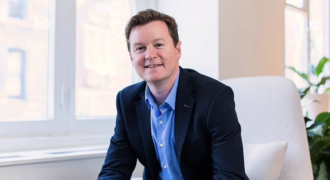 Colm O'Cuinneain, managing director of Greenhouse EMEA, is sitting in a modern, bright office, wearing a suit and smiling into the camera.