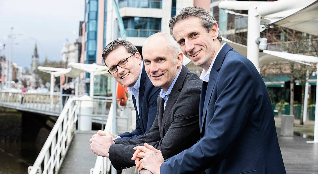 Three professional men from Granite Digital are standing in a row along a river bank in an industrial setting, smiling into the camera.