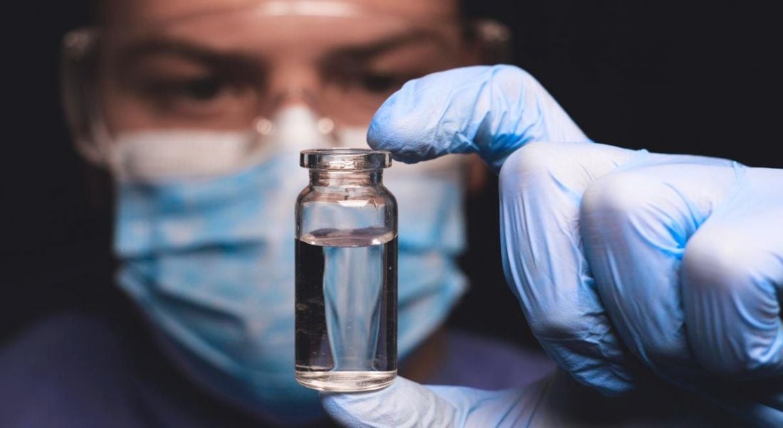 A biotech scientist is looking into a vial containing clear liquid while wearing lab gloves.