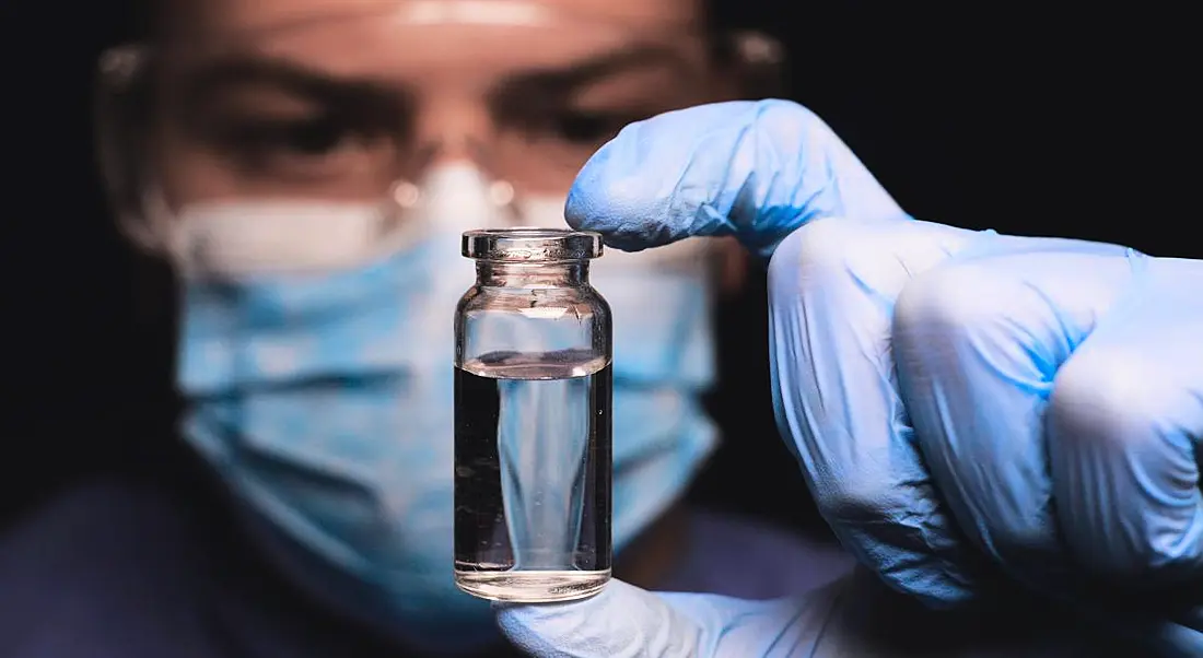 A biotech scientist is looking into a vial containing clear liquid while wearing lab gloves.