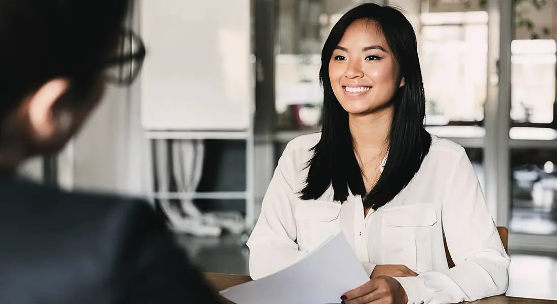 A professional woman is smiling at an interviewer across a table in a corporate office setting.