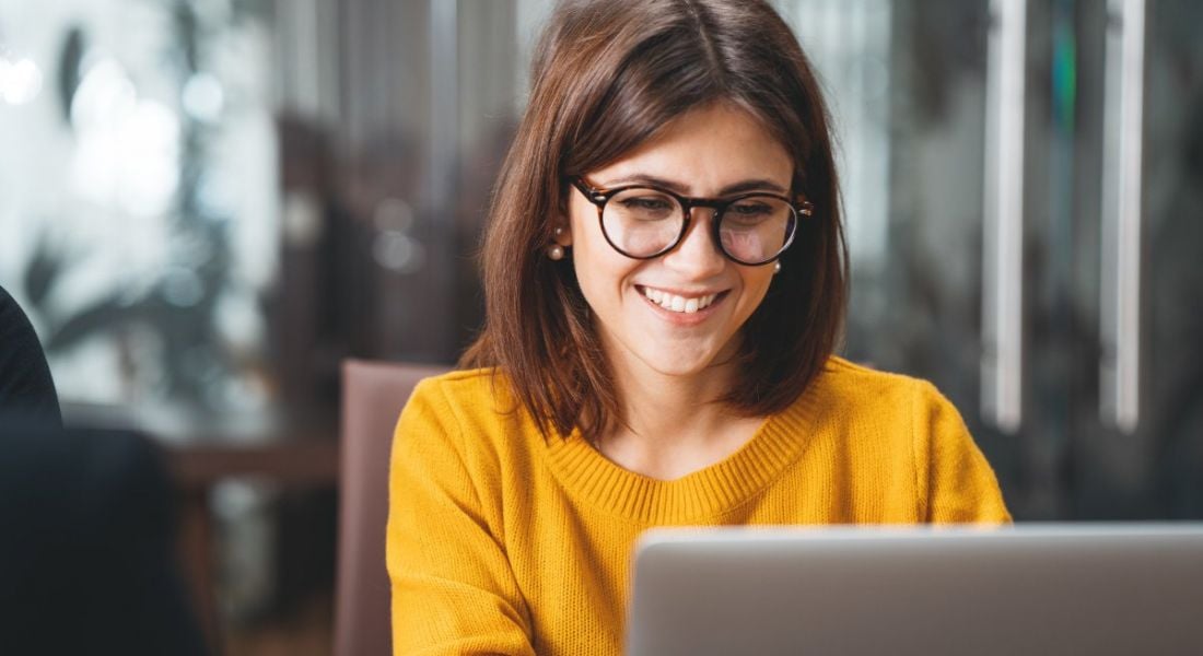 A happy woman wearing glasses and a yellow jumper is working at a laptop in an office.
