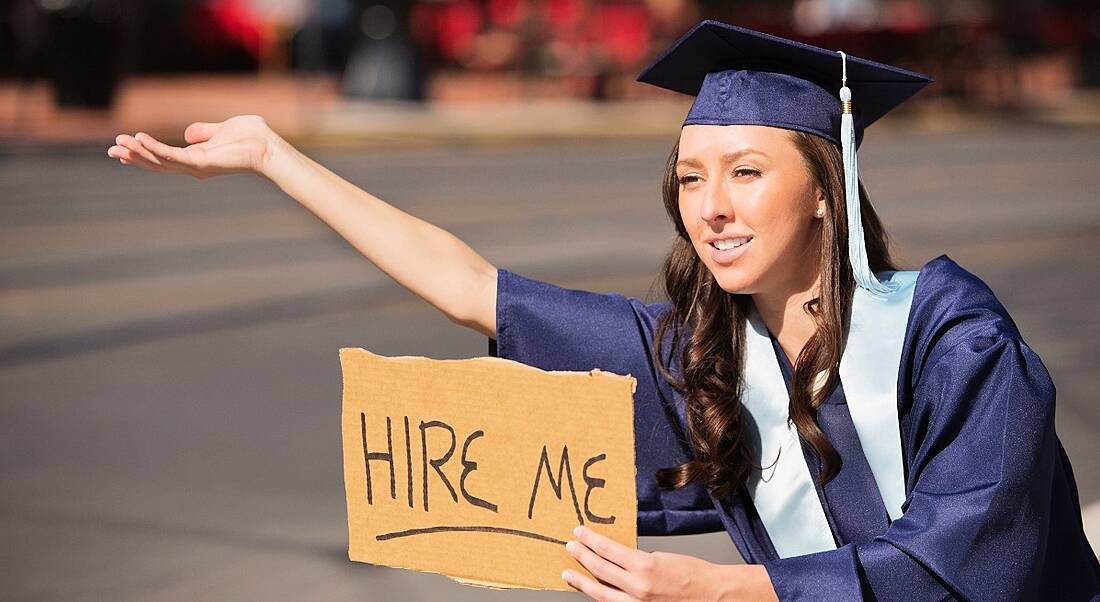 College graduate in gown holding a 'hire me' sign on the side of a road, searching for a graduate job.