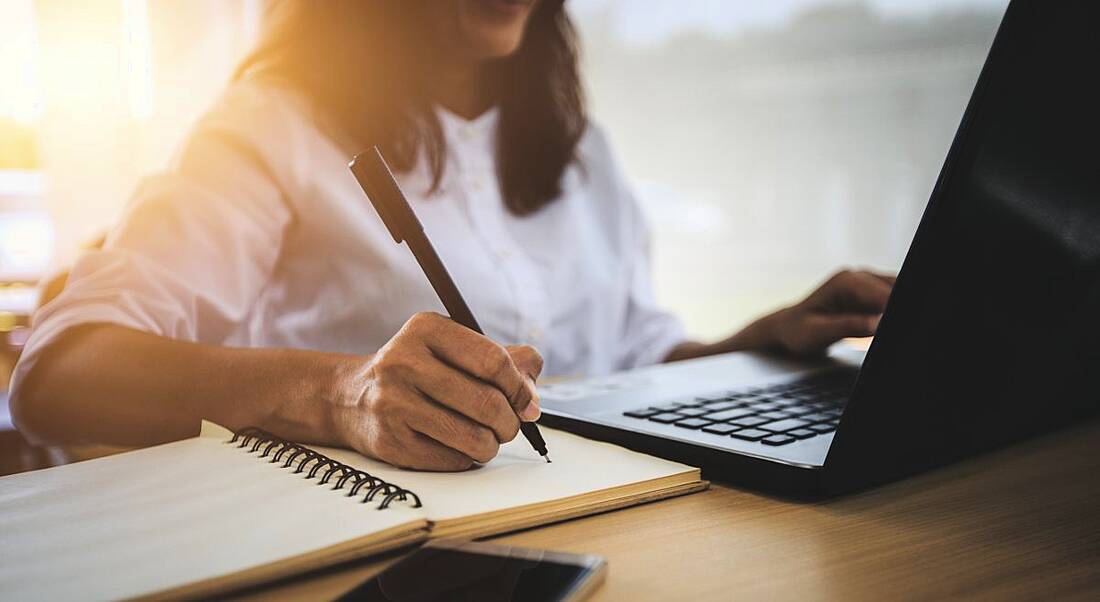 A woman is using a laptop and writing in a notepad at an office desk.