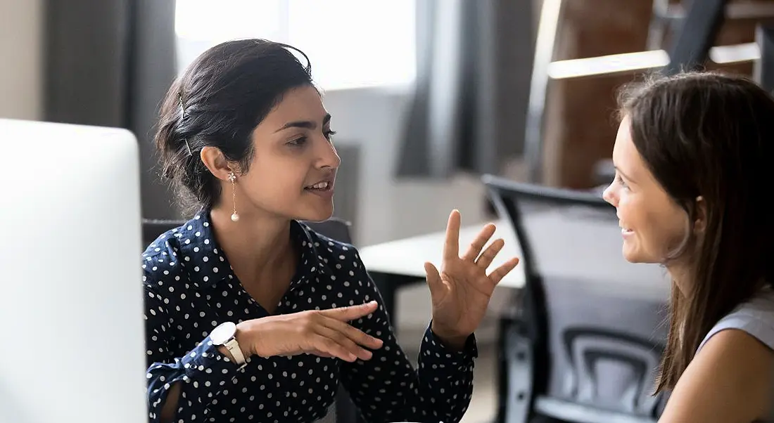 Two professional women are talking animatedly in an office setting, symbolising a good mentor and mentee relationship.