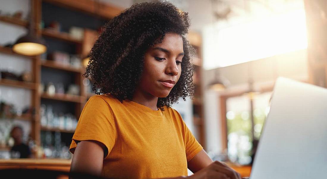 Young woman wearing orange T-shirt sitting in cafe working on her laptop.