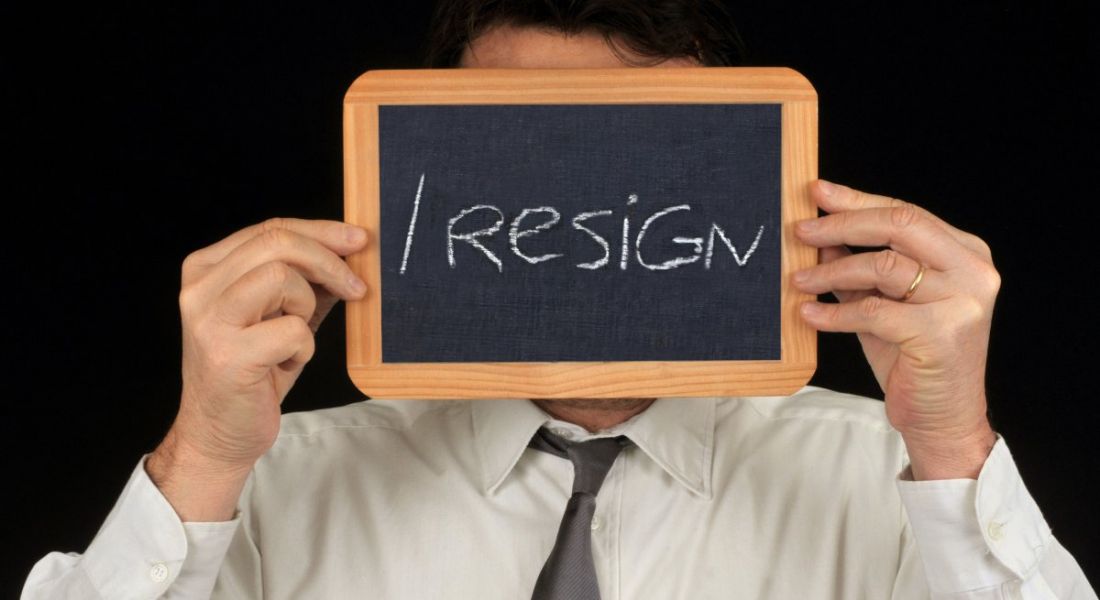 A man in business attire is standing against a black background and holding up a mini blackboard that says he is handing in his resignation.