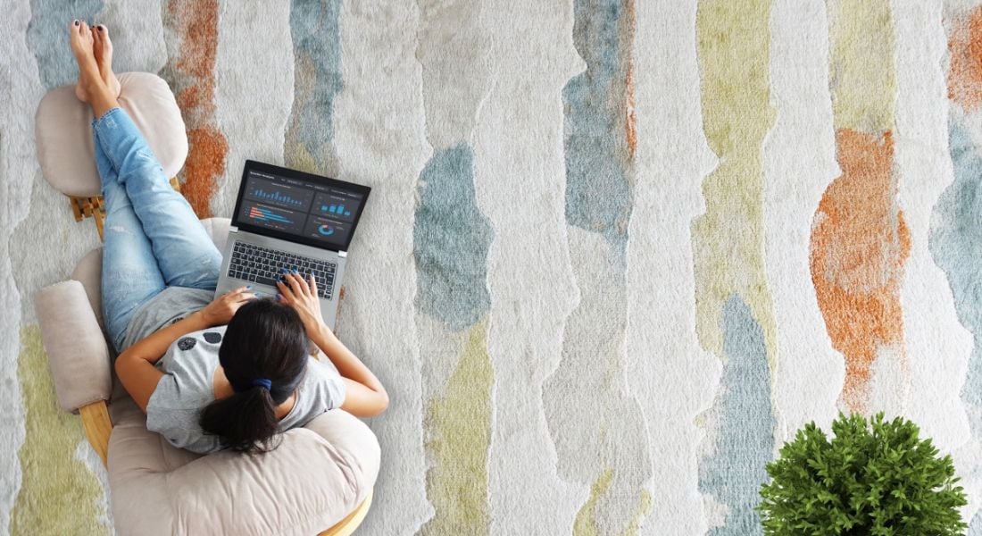 Woman remote working using a laptop from a comfortable chair, which is on a colourful carpet.