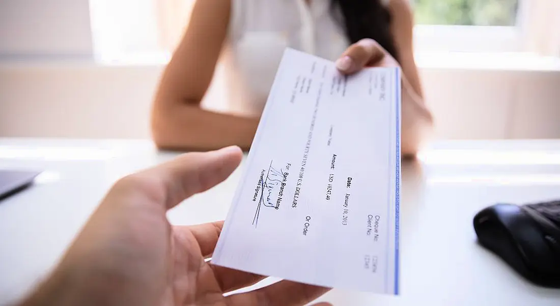 A person is handing a colleague their salary cheque across an office desk.