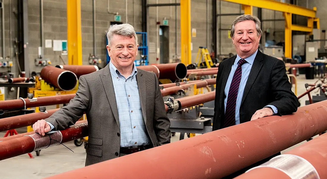 Two men in suits are smiling into the camera in a manufacturing room with large pipes.