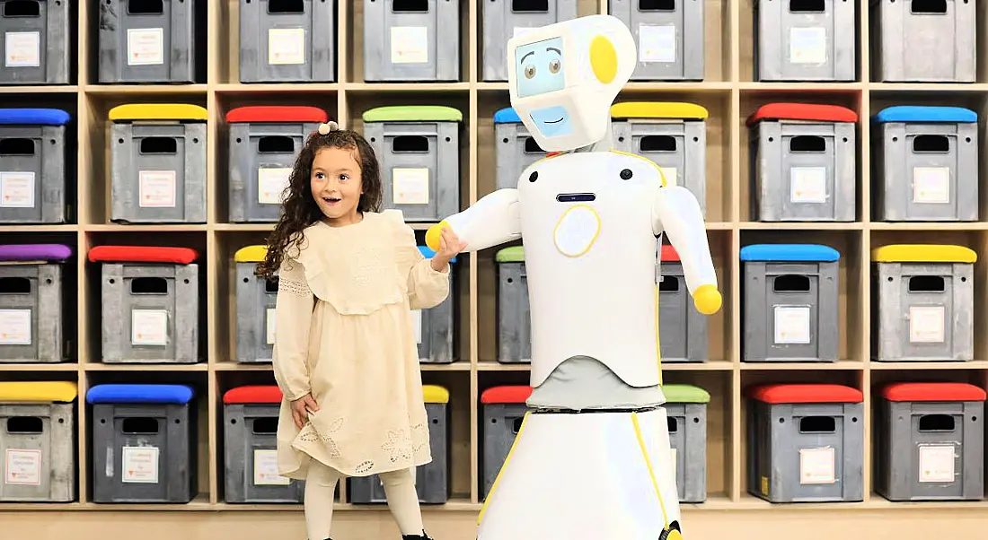 For Engineers Week, a young girl with dark hair excitedly holds the hand of a white and yellow robot, who is smiling.