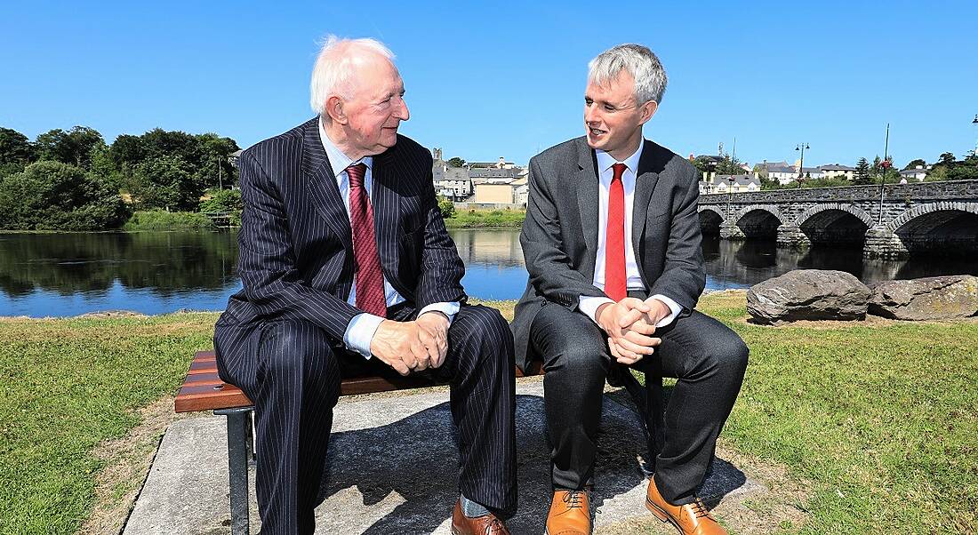 Brian McCarthy and Denis McCarthy sitting on a stone bench chatting in suits with a river and bridge behind them.