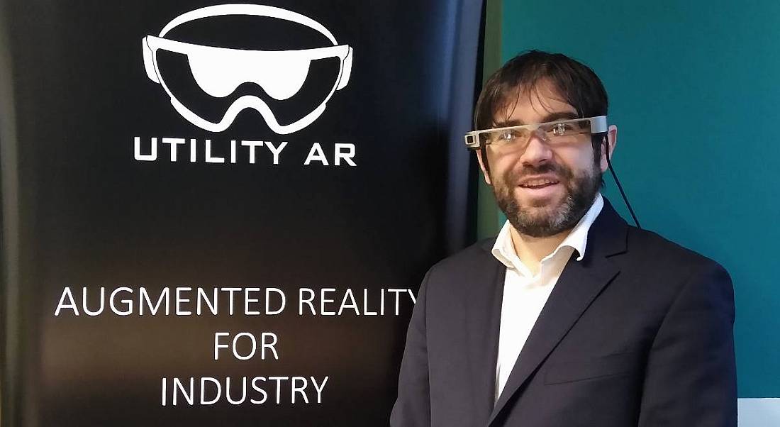 Patrick Liddy of UtilityAR is wearing AR glasses and smiling into the camera beside a pop-up stand with the company's branding on it.