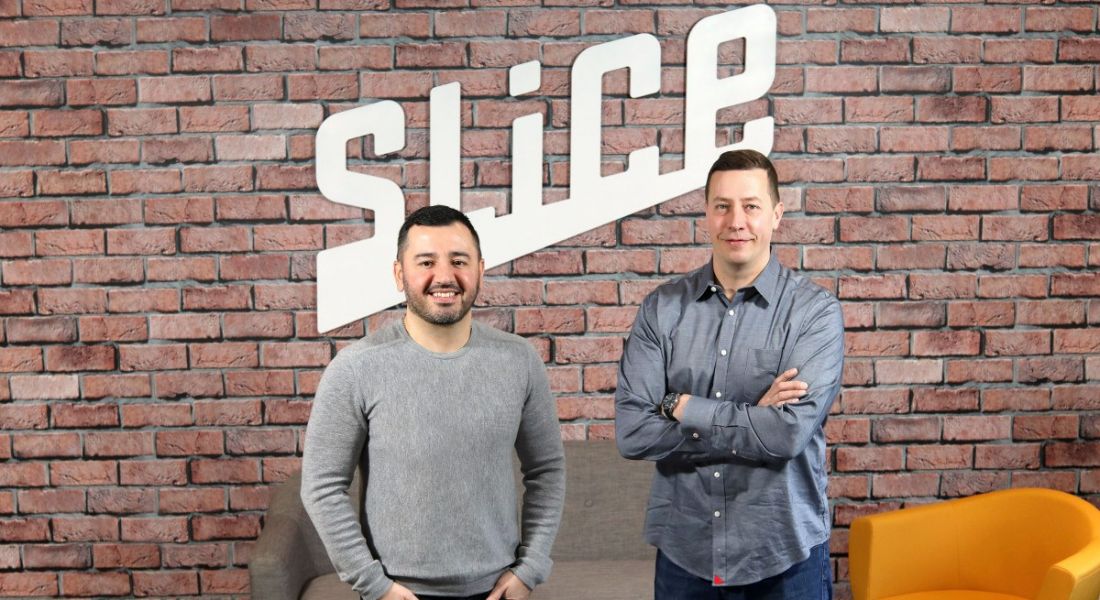 Two Slice employees are standing against a brick wall with Slice written on it, smiling into the camera.