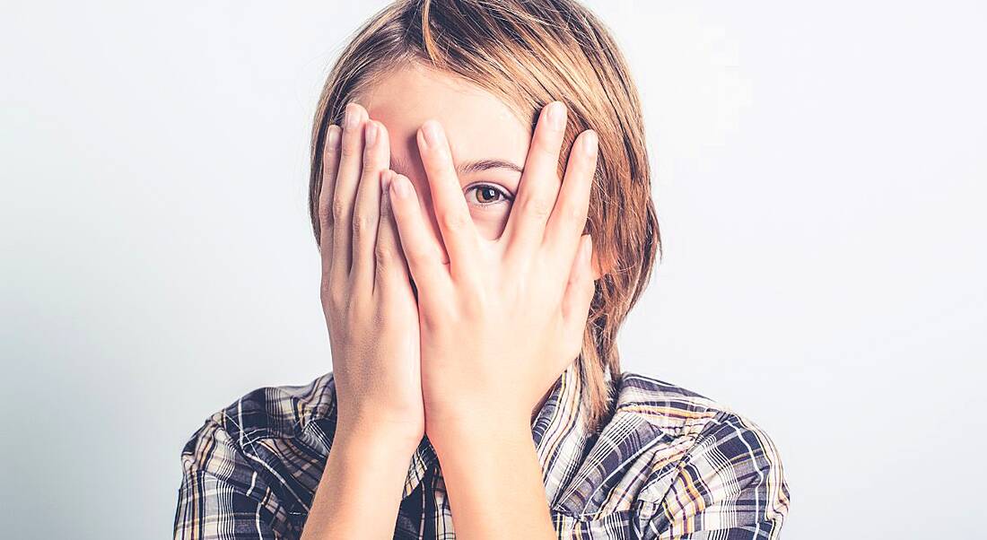 A young boy is holding his hands up to his face, peeking out from between his fingers in an introverted manner.