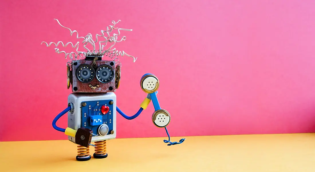 Colourful toy robot holding a telephone against a bright pink background.