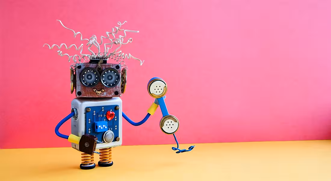 Colourful toy robot holding a telephone against a bright pink background.