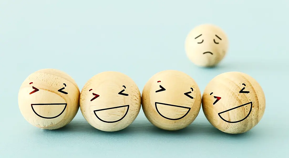 Four wooden balls with happy faces drawn on. A fifth one with a sad face is in the background, representing imposter syndrome.