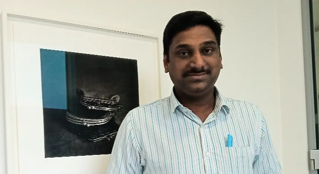 Kaarthik Janakiraman is standing in an office, wearing a striped shirt and smiling into the camera.