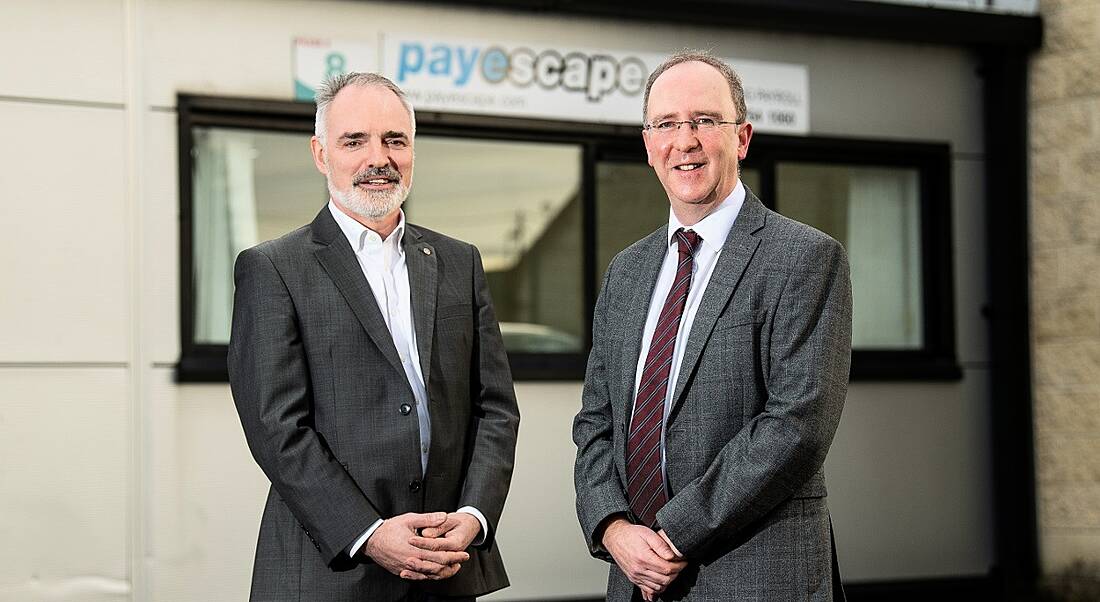 Two men in business attire are standing and smiling into the camera with a sign on a wall behind them for Payescape.