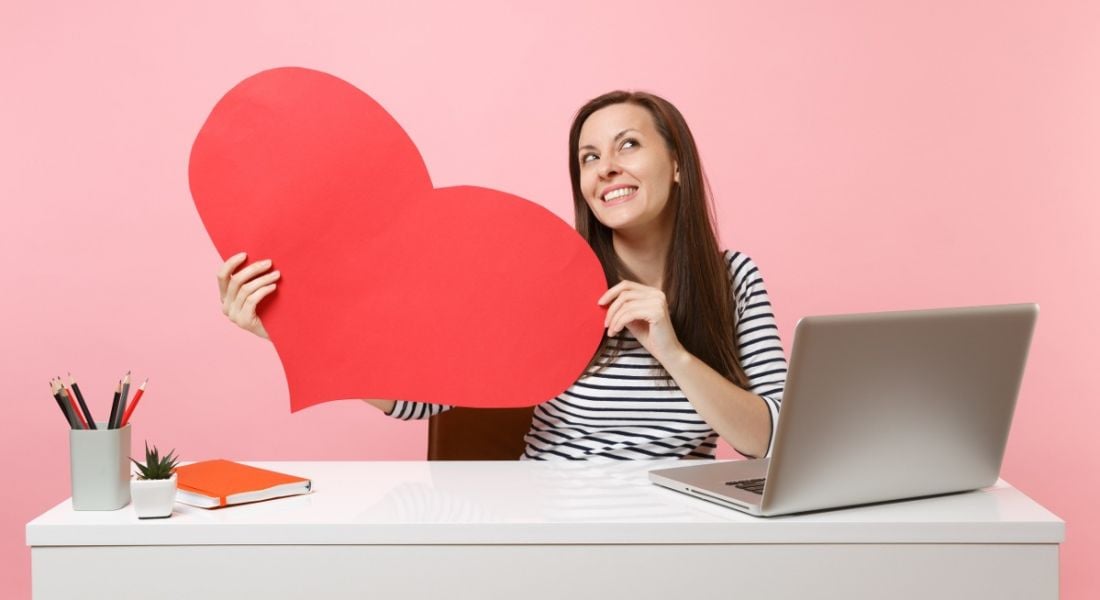 A woman is sitting at a desk with a laptop on it and is holding up a big pink paper heart.