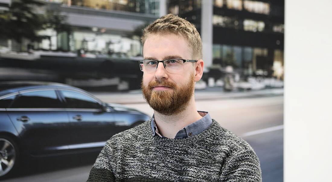 Aaron Hetherington is looking into the camera against a picture of a car on a street at the JLR offices in Shannon.