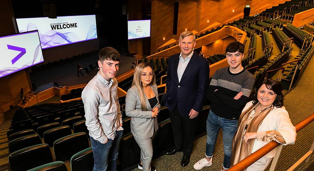 Three men and two women stand at the top of an auditorium with the stage in the background. The screen says welcome, with the Accenture logo.