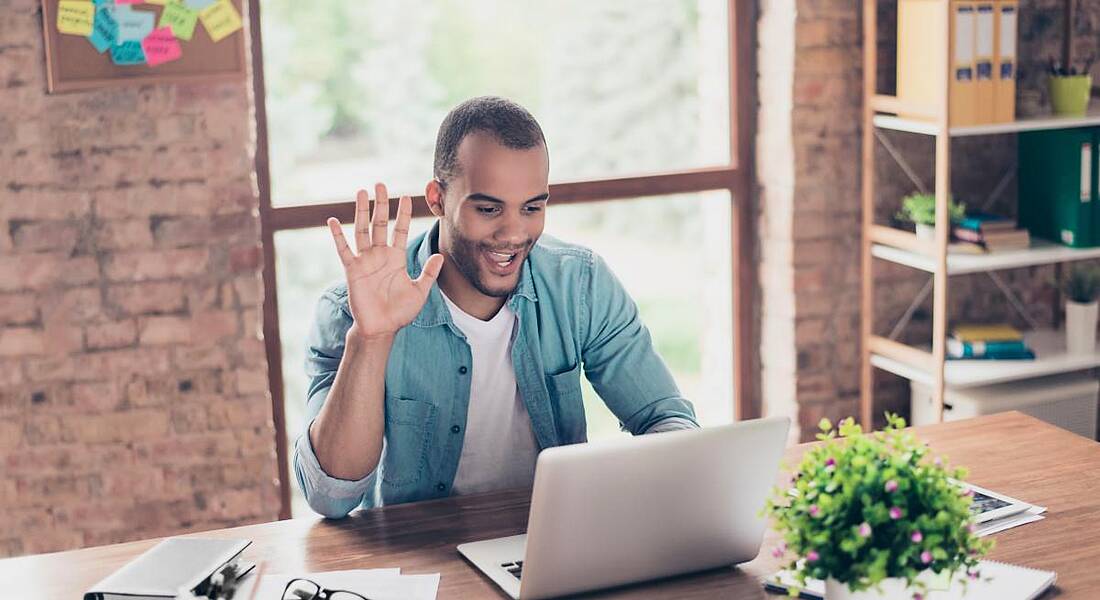 Cheerful employee is waving at the camera on his laptop while having a video call sitting at his work station in casual clothes.